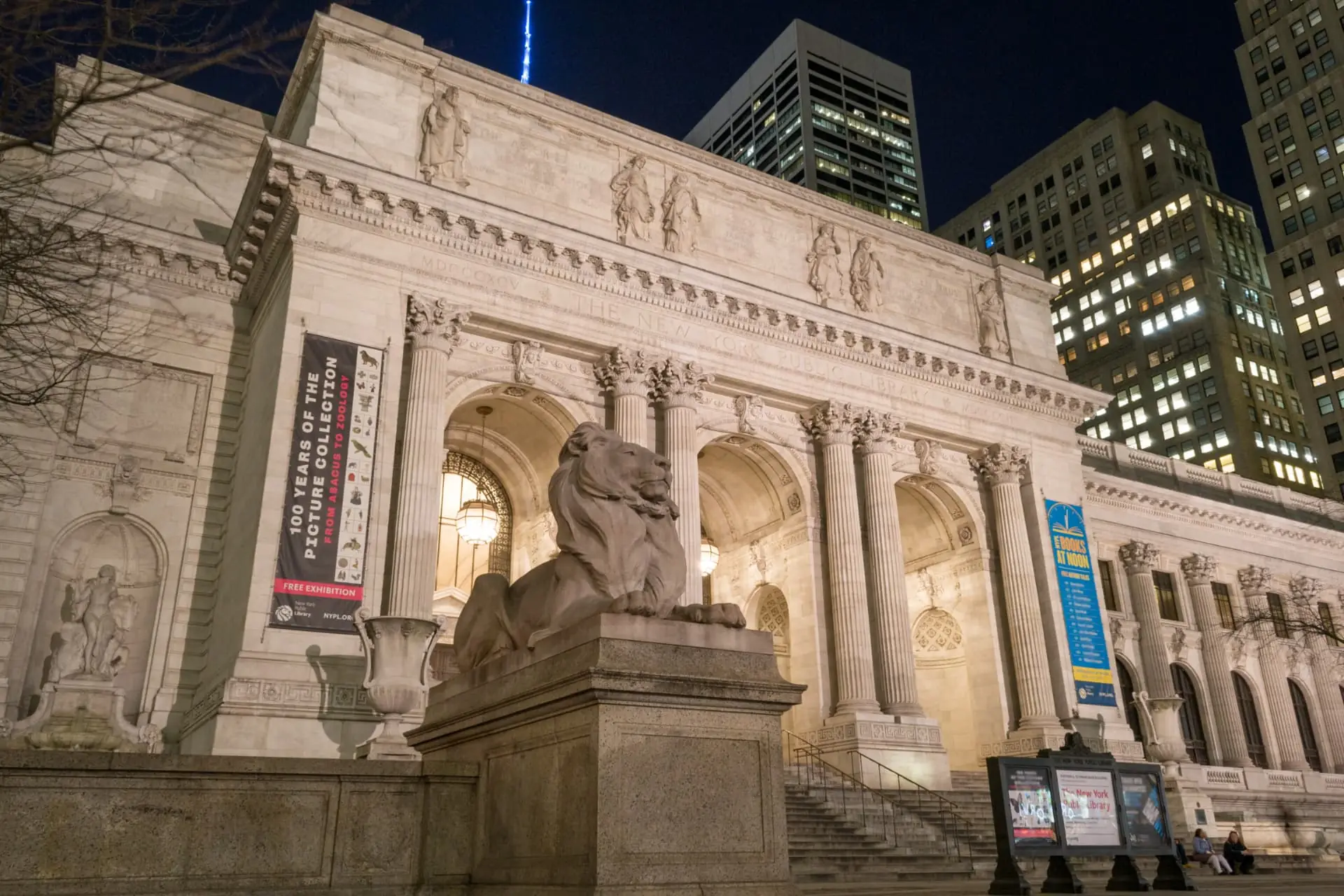 The New York Public Library lions lit at night with Midtown towers behind