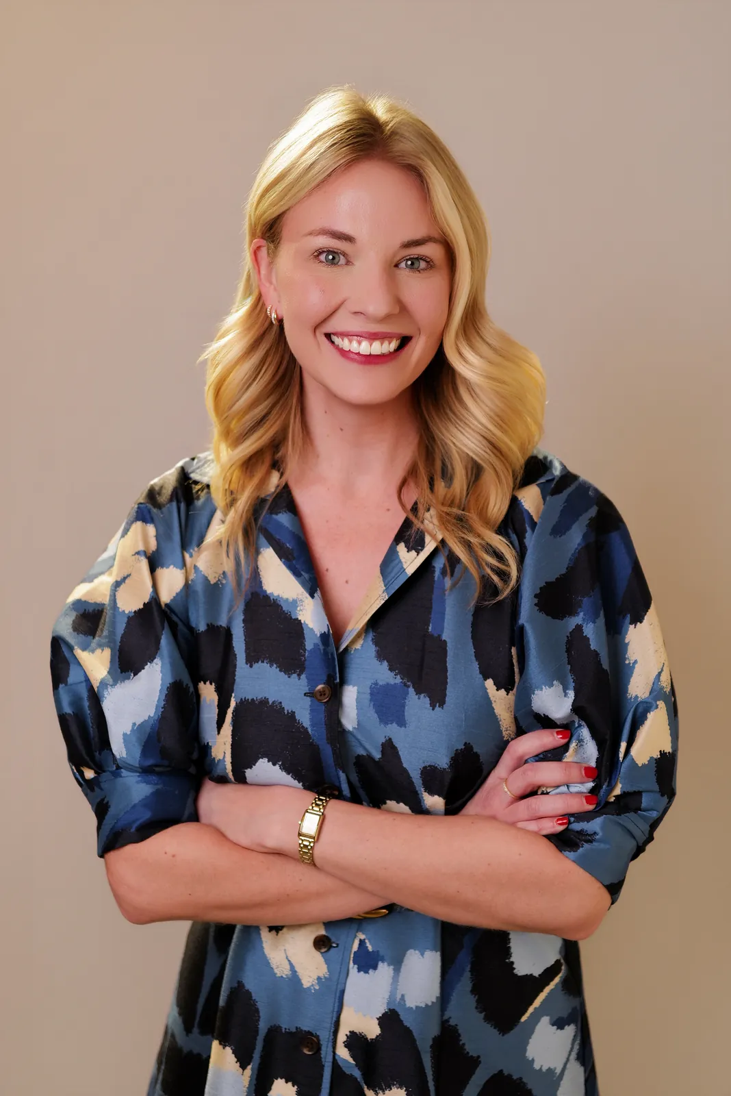 Studio headshot of a smiling blonde woman with arms crossed in a patterned blouse