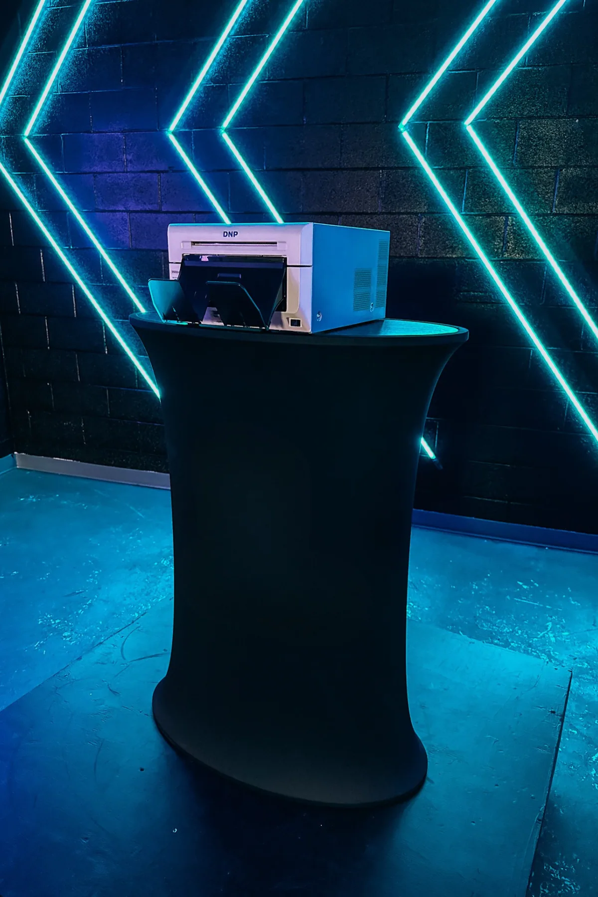 Unlimited printing station with DNP printer on a black cocktail table in front of teal neon chevron lighting